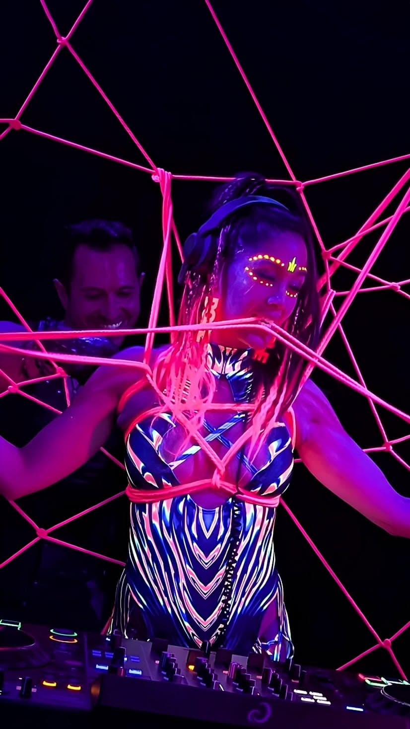 Aster at the decks inside a web of fluorescent rope under UV light, with Davide behind her, during a SUSPND performance.