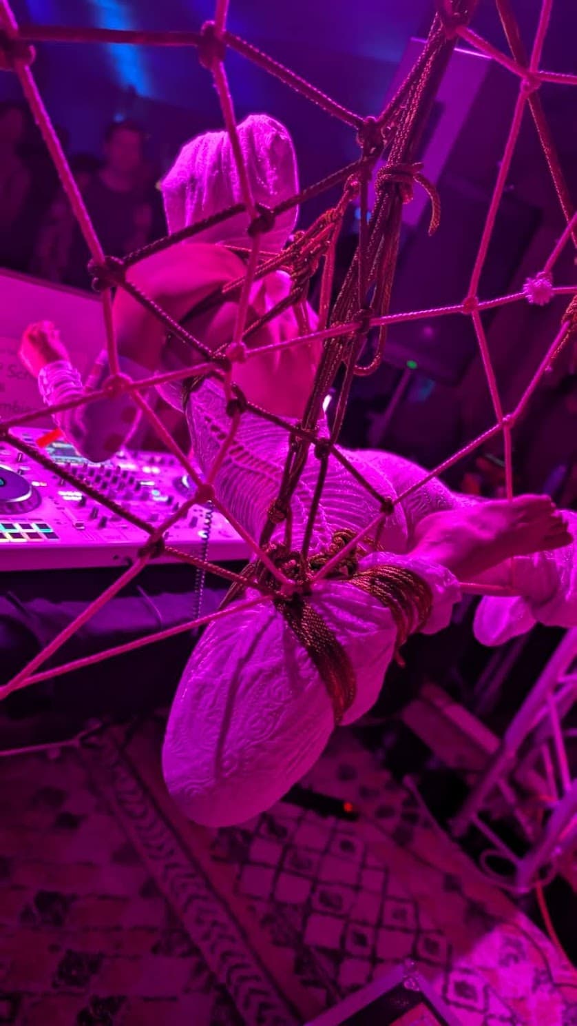 A full rope suspension held horizontally above the DJ booth during a SUSPND performance, lit in magenta.